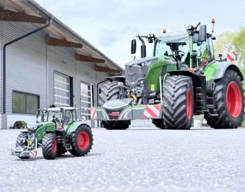 A large green tractor with a front weight bumper is parked outside a building next to a toy tractor with a similar bumper