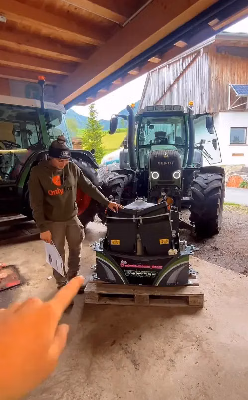 A person stands next to a Fendt tractor and a front weight bumper with LED lights resting on a pallet in a garage.