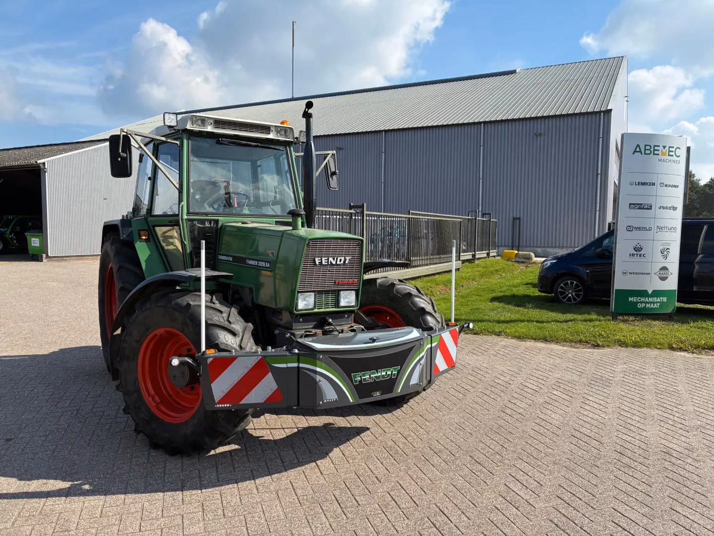 A green tractor with a front weight bumper is parked on a paved area near a building and a sign with company logos