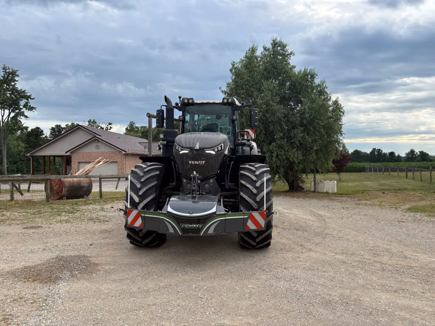 A large black tractor with a front weight bumper and LED lights is parked on a gravel driveway near a house and trees