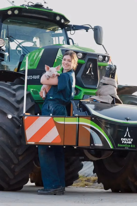 A person in overalls holding a piglet stands by a green tractor with a Deutz-Fahr front weight bumper attached