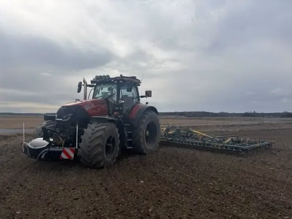 Red tractor with a front weight bumper and lights attached, working in a large open field with farm equipment behind
