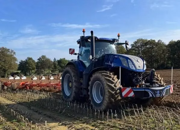 Blue tractor with a front weight bumper attached, working in a field with a plough implement in the background