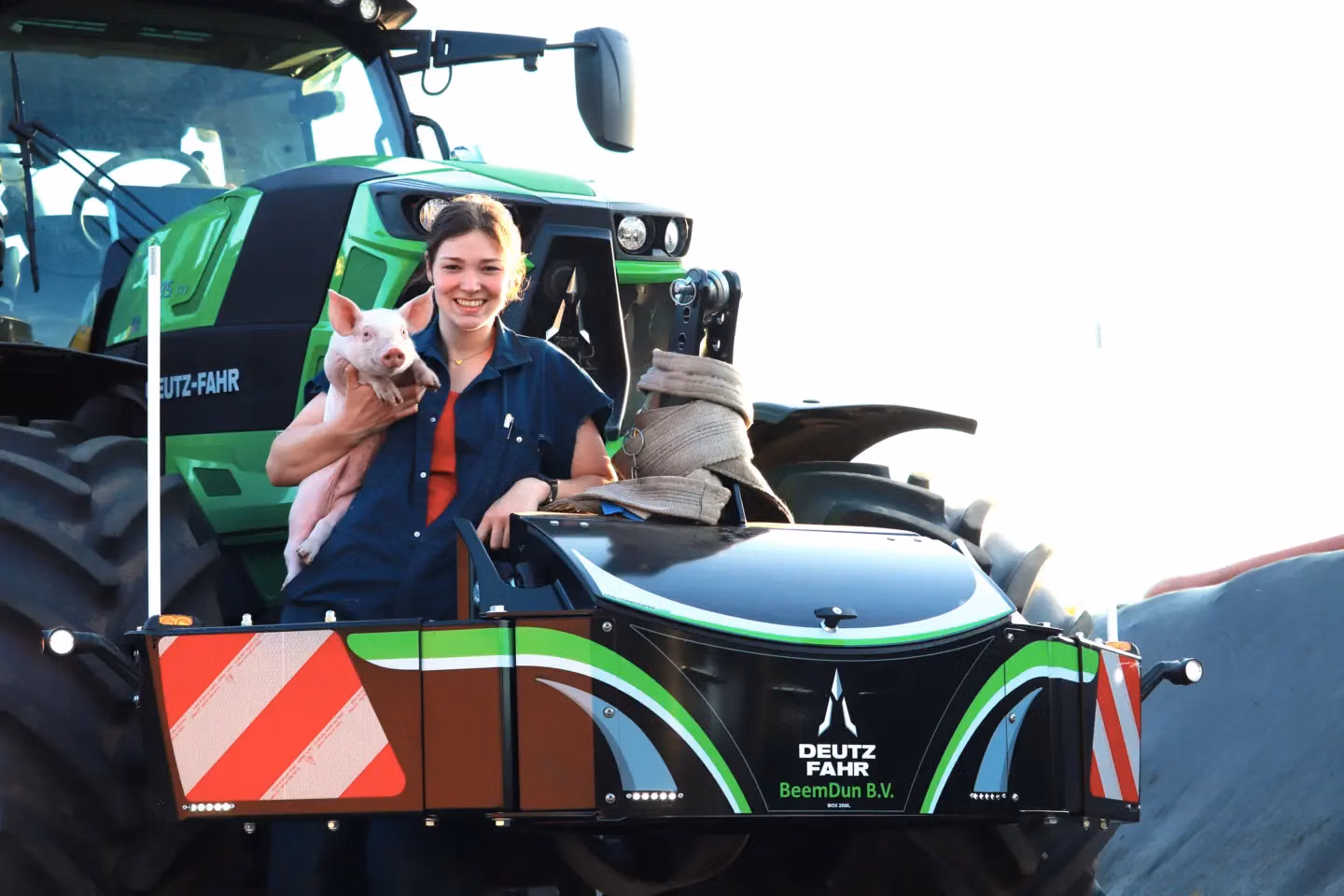 Smiling person holding a piglet stands by a Deutz-Fahr front weight bumper attached to a green tractor.