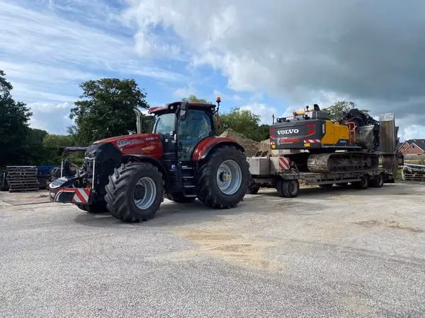 A red tractor fitted with a front weight bumper is towing a flatbed trailer carrying a tracked excavator.