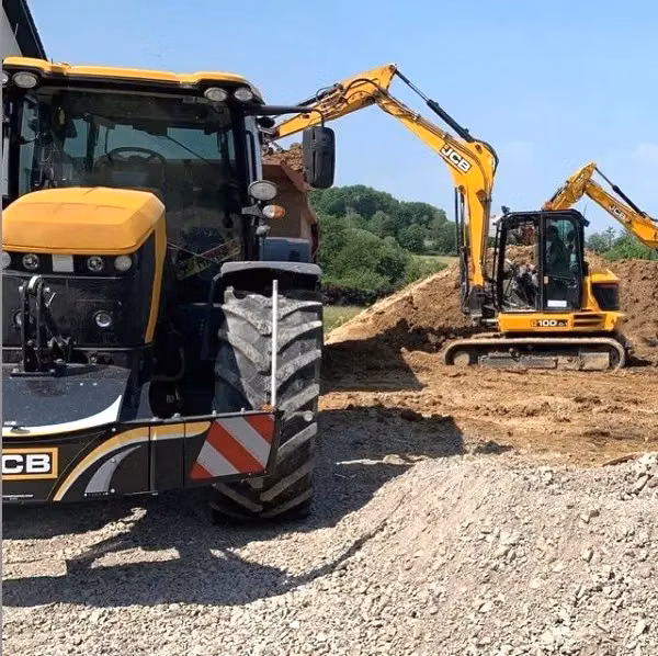 A tractor with a front weight bumper is parked on gravel near two excavators working on a dirt mound in the background