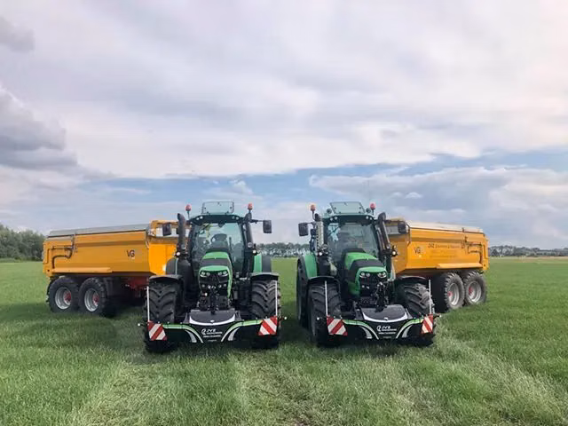 Two green tractors with front weight bumpers are parked side by side on grass, each attached to a yellow trailer