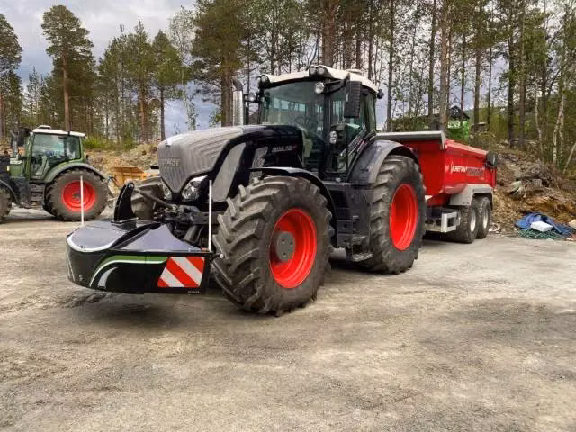 Black tractor with a front weight bumper attached, pulling a red trailer, parked outdoors on a dirt surface