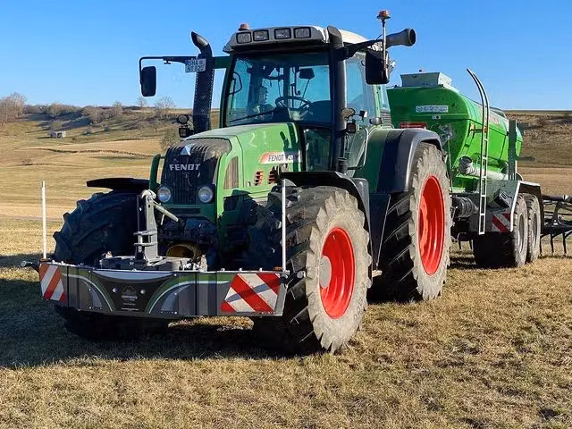 Green tractor with front weight bumper and red wheels hitched to a green agricultural trailer on grassy terrain
