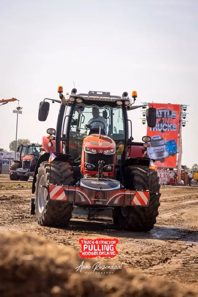 Red tractor with a front weight bumper on a dirt track at an outdoor event with people and banners in the background