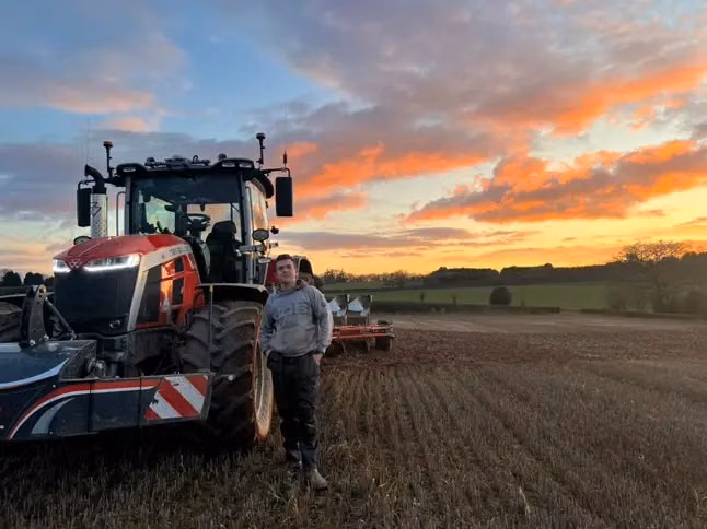 A person stands in front of a tractor with a front weight bumper and LED box in a field at sunset