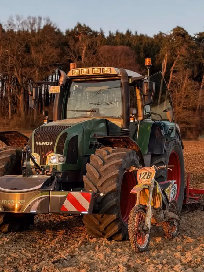 Green tractor with a front weight bumper attached, parked in a field beside a dirt bike with number 126