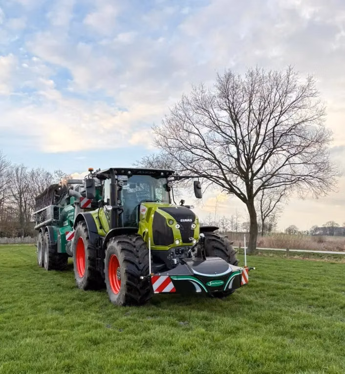 Green tractor with a front weight bumper attachment parked on grass beside a leafless tree under a partly cloudy sky