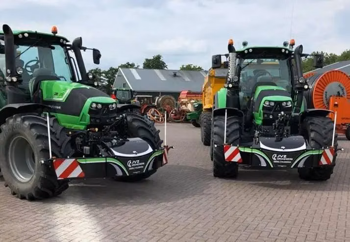 Two green tractors parked on pavement, each fitted with a black front weight bumper with red and white stripes