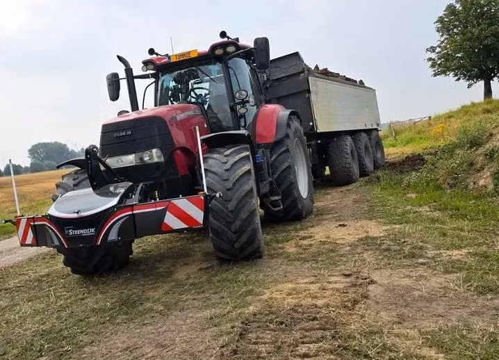 A red tractor with a front weight bumper is towing a large trailer filled with loose material on a grassy path