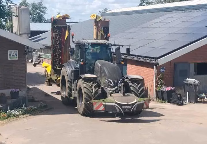 A black tractor with a front weight bumper is parked on a farmyard between brick buildings and flower pots