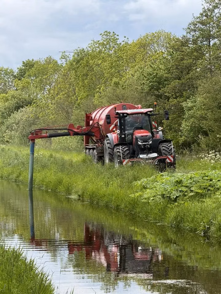 Red tractor with a front weight bumper and large tank attachment parked beside a grassy canal with trees in the background