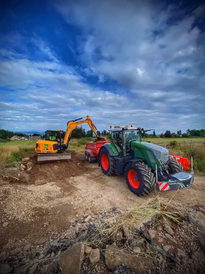 Green tractor with a front weight bumper attached, next to an excavator and trailer on a gravel area outdoors