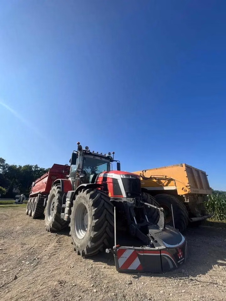 Large tractor with a front weight bumper attachment parked on a dirt surface next to a yellow trailer under clear blue sky