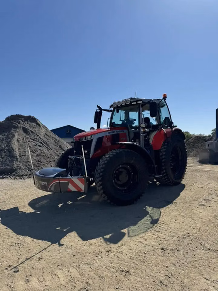 Red tractor with a front weight bumper attachment parked on a dirt surface near piles of gravel