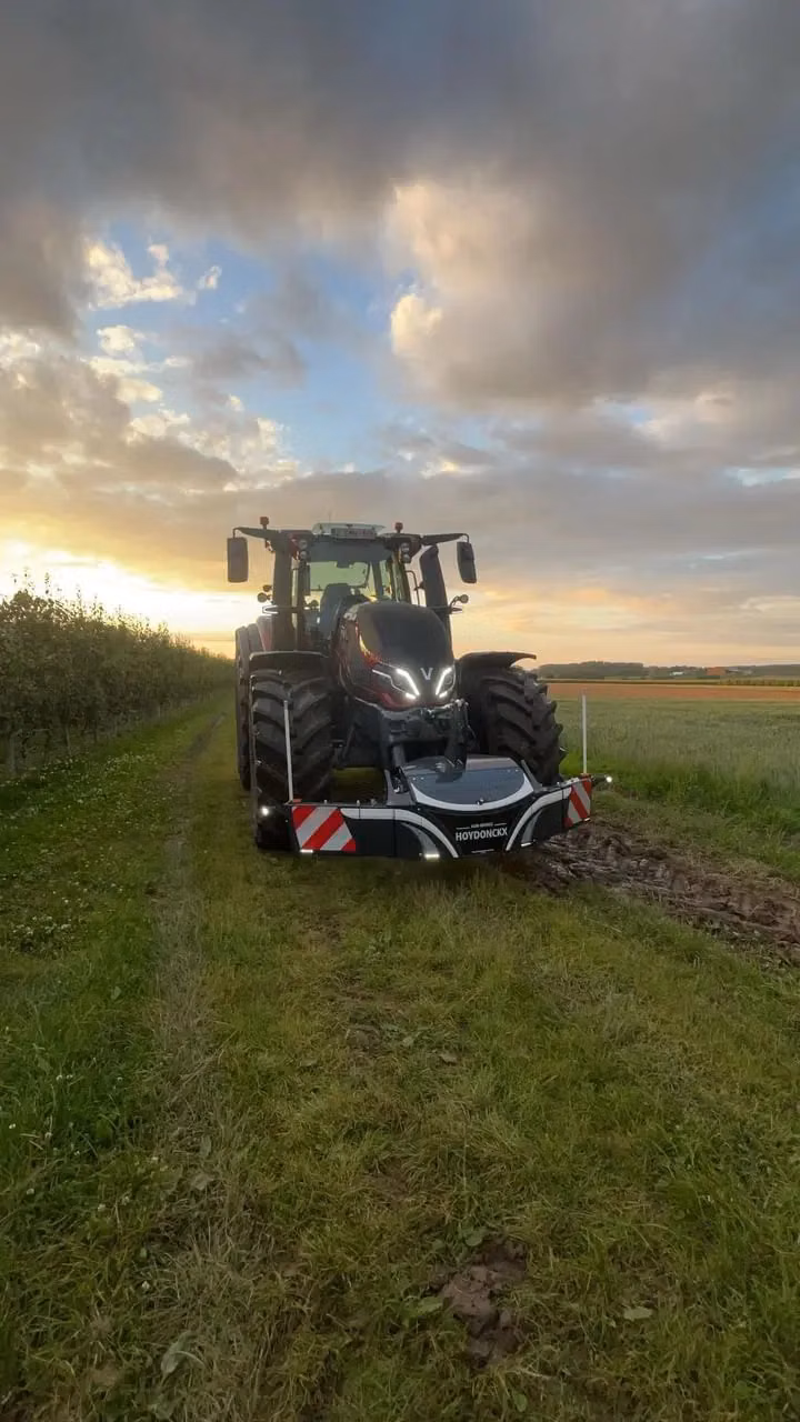 A tractor with a front weight bumper labeled HOYDONCKX is parked on a grassy path beside a field at sunset