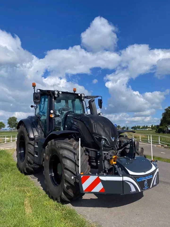 Large black tractor with oversized tires fitted with a front weight bumper on a road beside green grass under a blue sky