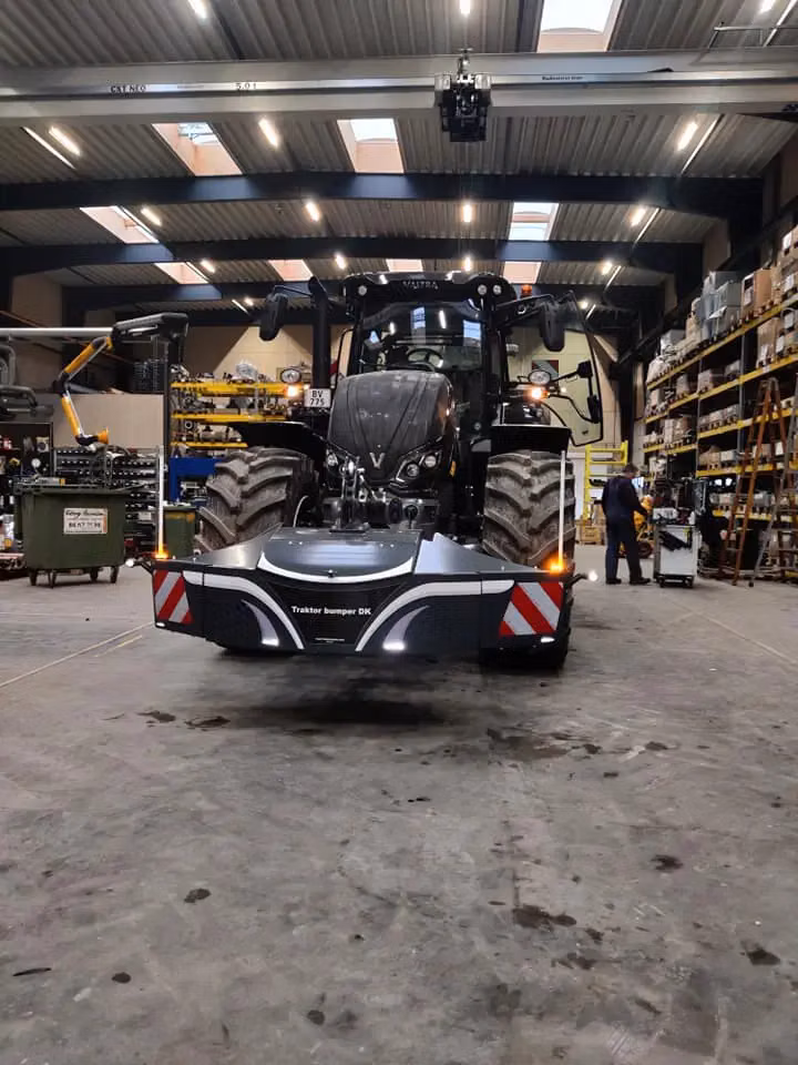 Tractor in a workshop fitted with a front weight bumper displaying reflective panels and labeled Traktor bumper DK