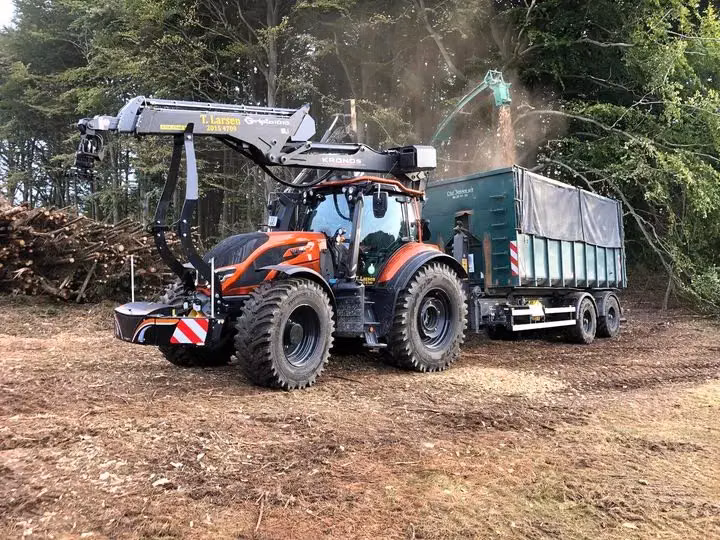 Orange tractor with front weight bumper, loader crane and attached trailer collecting wood chips in a forest clearing