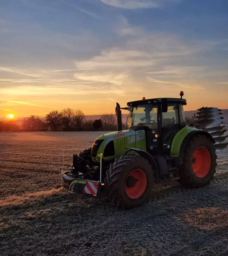 A tractor with a front weight bumper is parked on a frosty field at sunrise with a plough attached at the rear