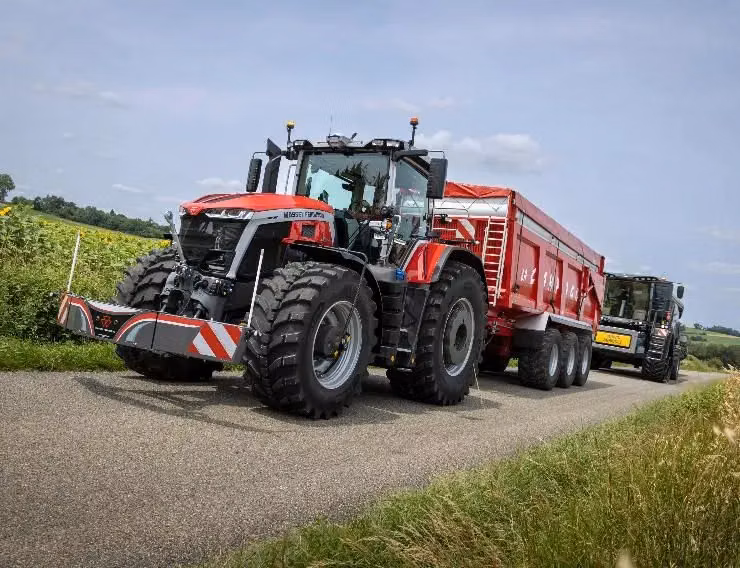 Red tractor with a front weight bumper and large trailer attached, driving on a rural paved road