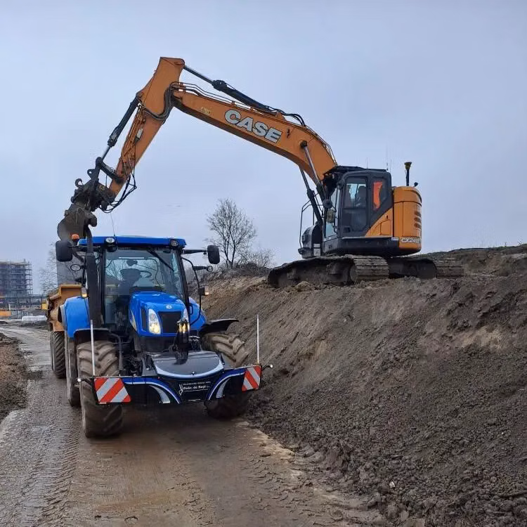 A blue tractor with a front weight bumper is parked on a dirt road while an excavator loads its trailer
