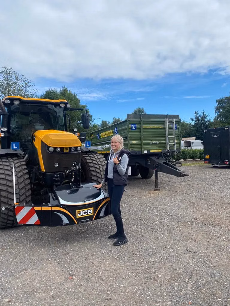 A woman stands beside a yellow tractor with a front weight bumper labeled JCB and a green trailer in the background