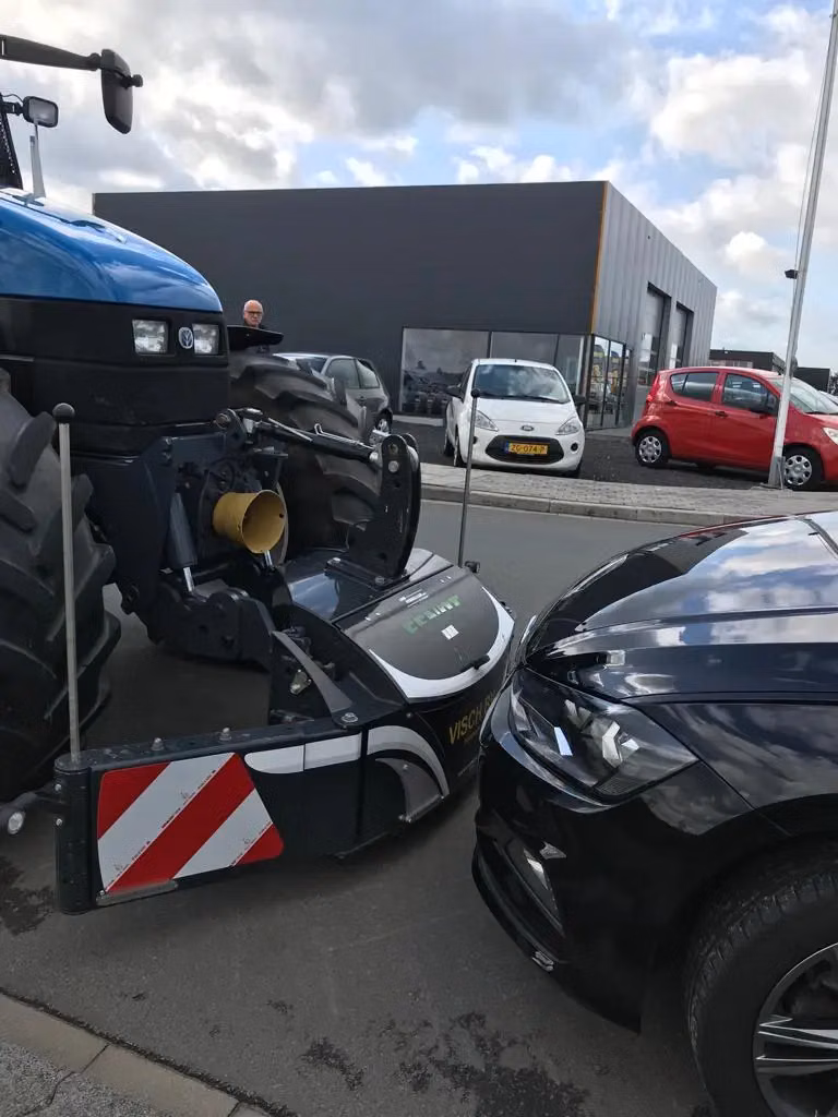 A blue tractor with a front weight bumper is positioned closely in front of a black car on a street.
