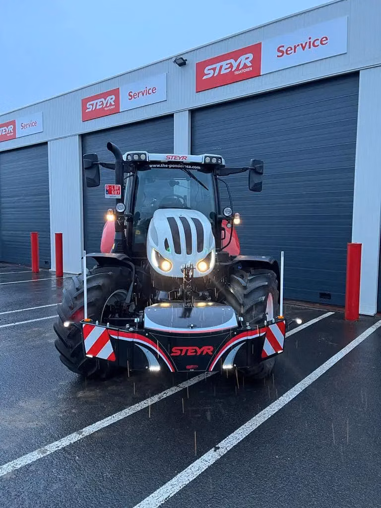 Tractor with headlights on, fitted with a front weight bumper labeled STEYR, parked in front of service garage doors