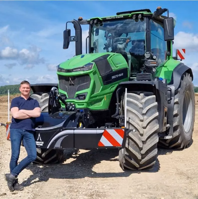 A person stands beside a green tractor with a front weight bumper featuring red and white reflective stripes