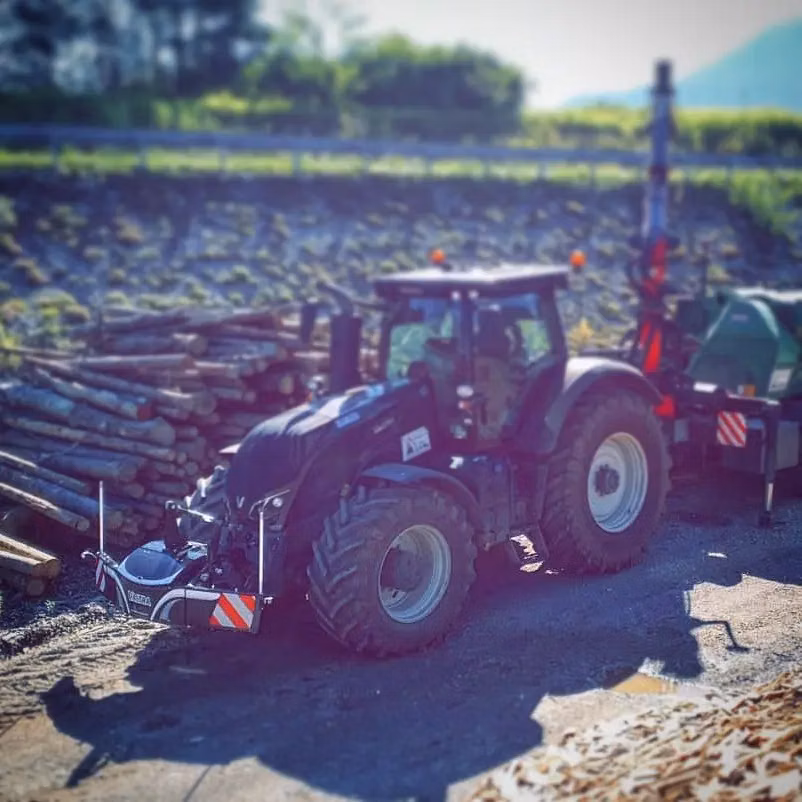 A tractor with a front weight bumper is parked beside stacked logs and attached to a trailer in a rural area