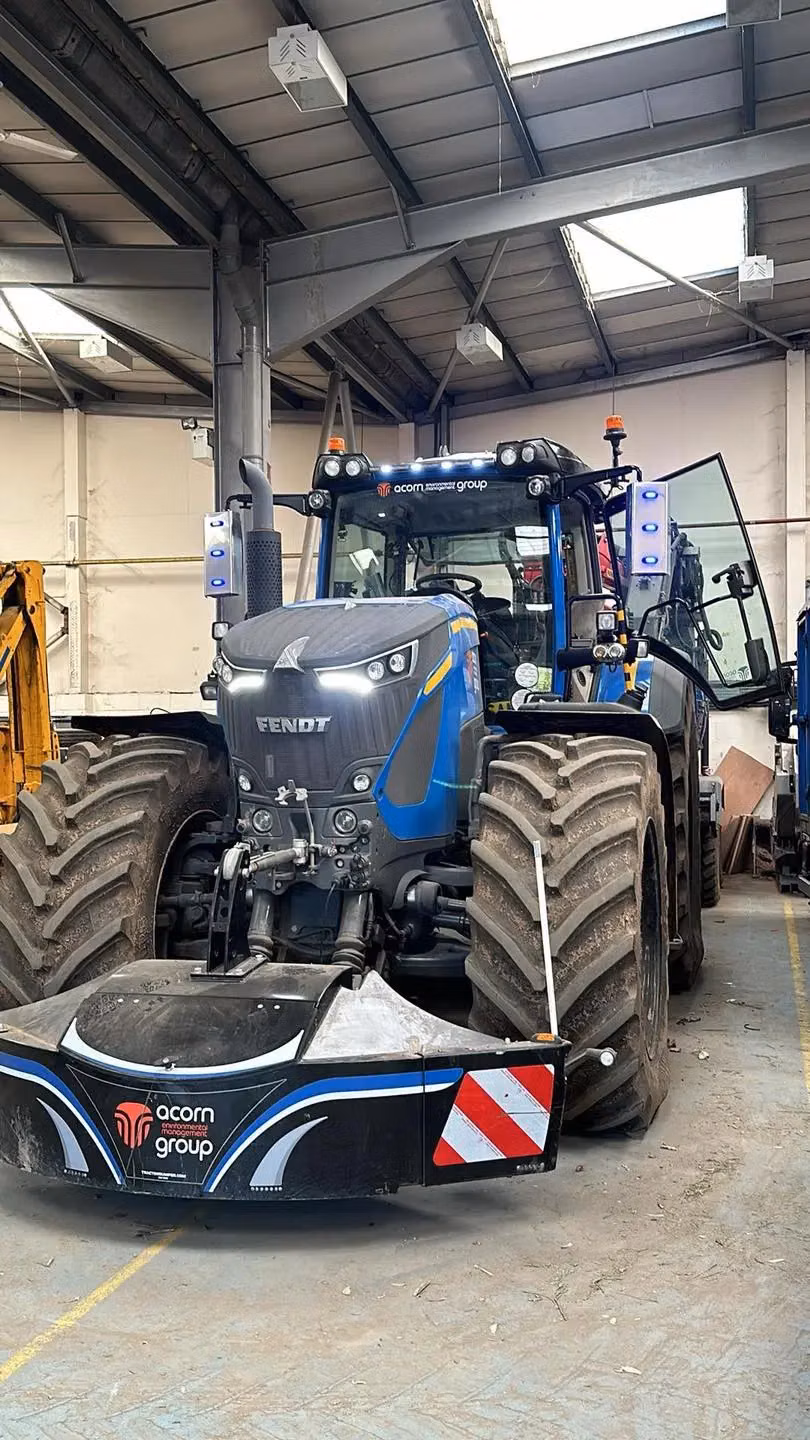 A large blue tractor indoors fitted with a front weight bumper featuring lights, safety markings and acorn branding