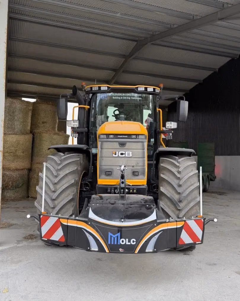 Large tractor with a MOLC branded front weight bumper, parked indoors on concrete beside stacked hay bales