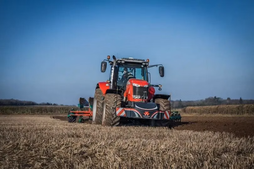 A red tractor with a front weight bumper is plowing a field under a clear blue sky