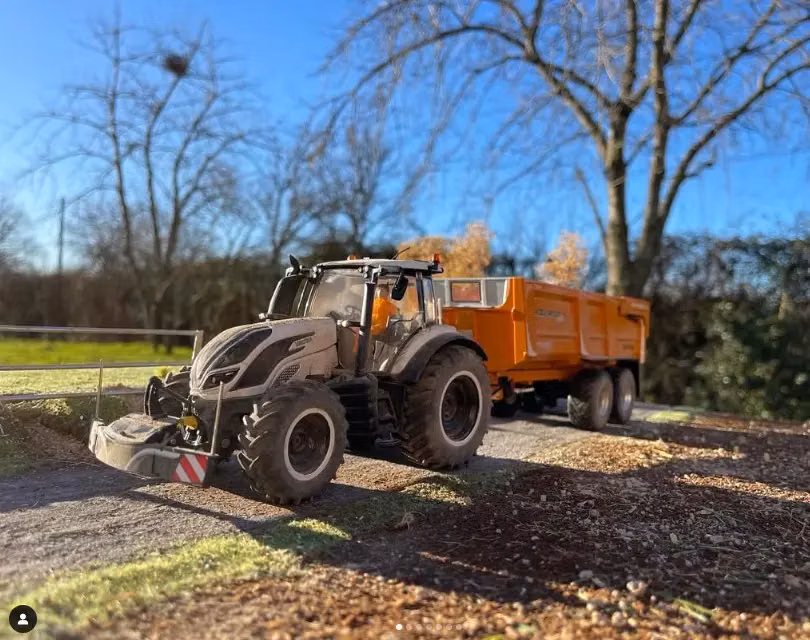 A tractor with a front weight bumper and an orange trailer is parked on a rural dirt path with trees in the background