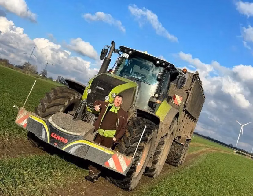 A person stands in front of a tractor with a CLAAS front weight bumper on a grassy field under a blue sky.