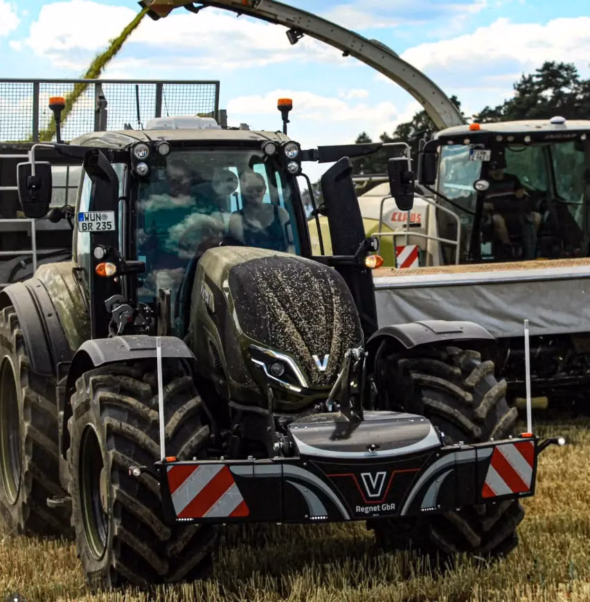 A tractor with a front weight bumper and LED lights works in a field near harvesting machinery and tall grass