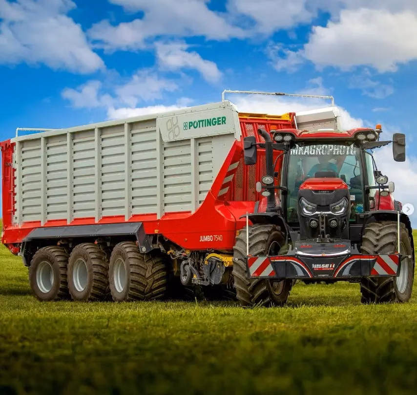 A red tractor with a front weight bumper is hitched to a large multi-axle forage wagon on grass under a blue sky