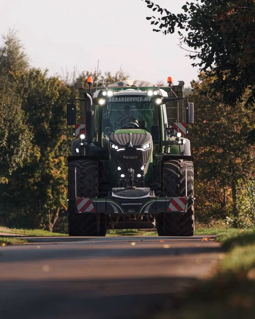 A green tractor with wide front weight bumper, LED lights and reflective markings on a rural road