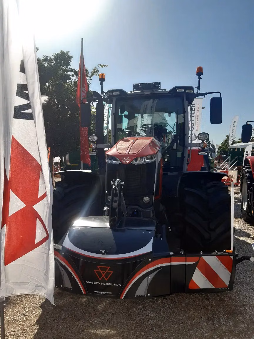 A red tractor with a Massey Ferguson front weight bumper displayed outdoors, with flags and signs around it