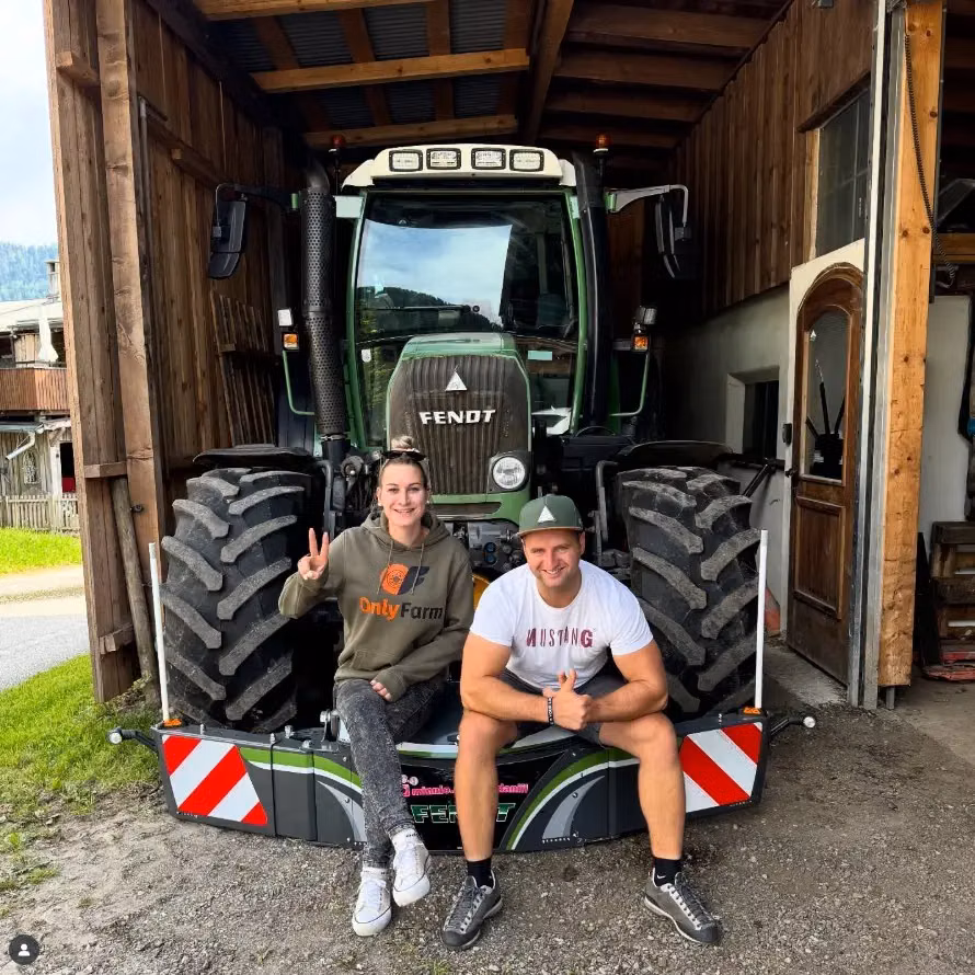 Two people sit on a front weight bumper with LED lights and reflective strips mounted on a Fendt tractor in a barn entrance