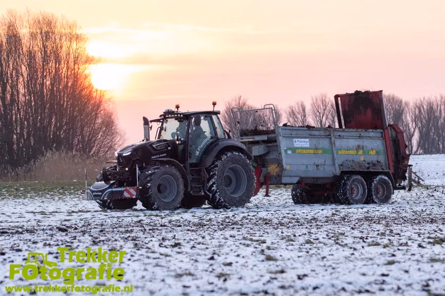 Black tractor with a front weight bumper attached, pulling a loaded trailer on a snowy field at sunset