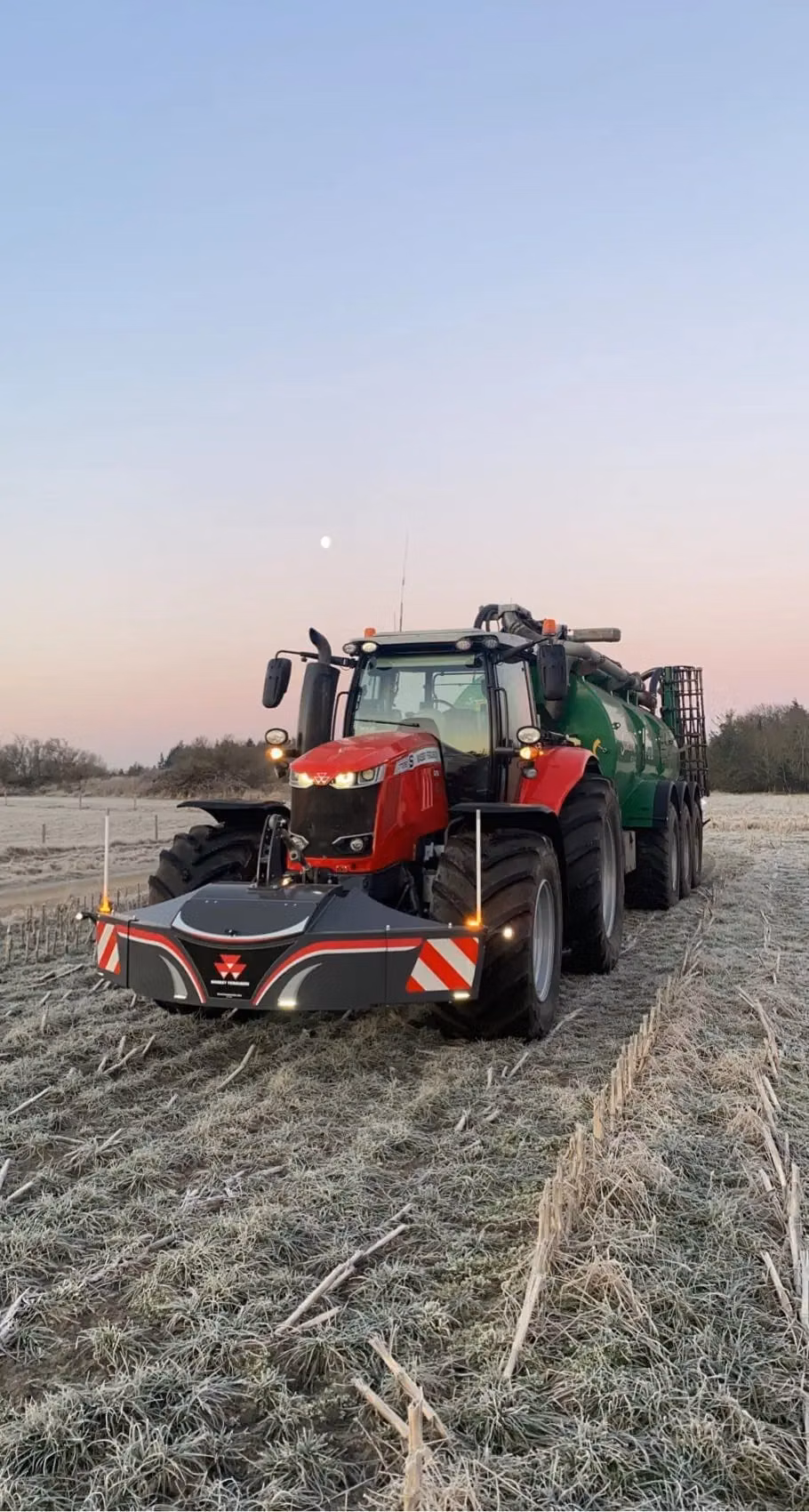 A red tractor fitted with a front weight bumper is parked on a frosty field at sunrise with a green trailer attached