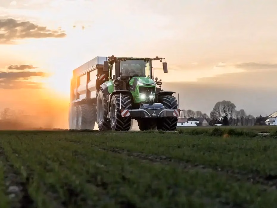 A green tractor with a front weight bumper is driving on a field at sunset with a trailer attached