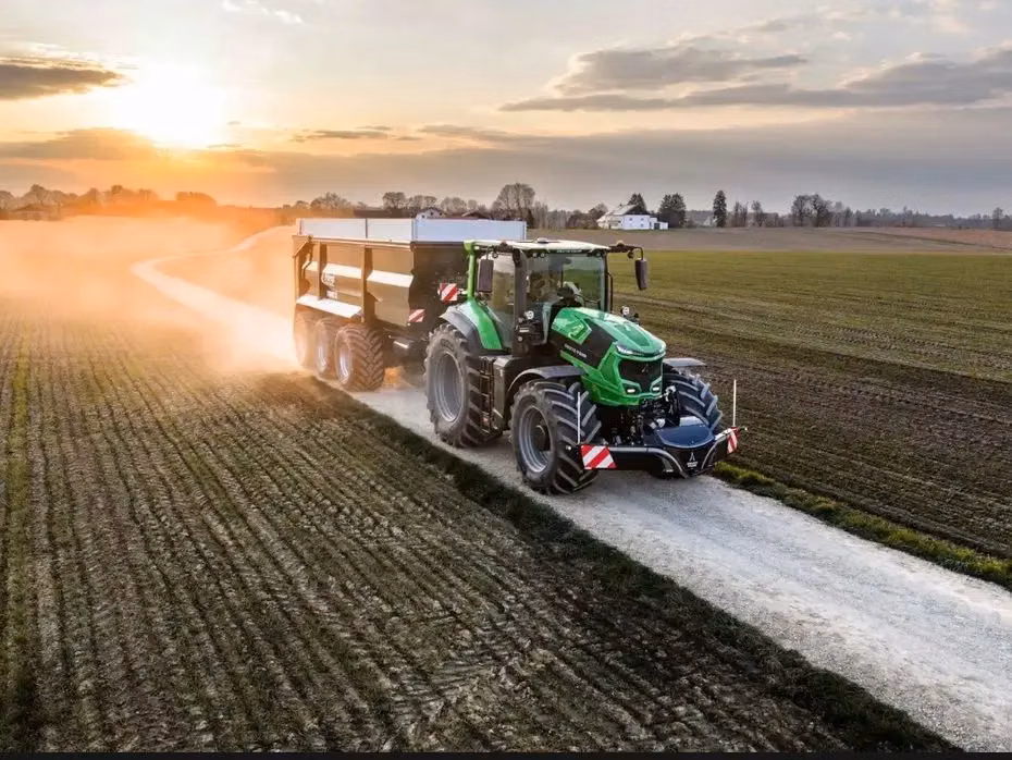 A green tractor with a front weight bumper and trailer drives on a dirt road beside farm fields at sunset